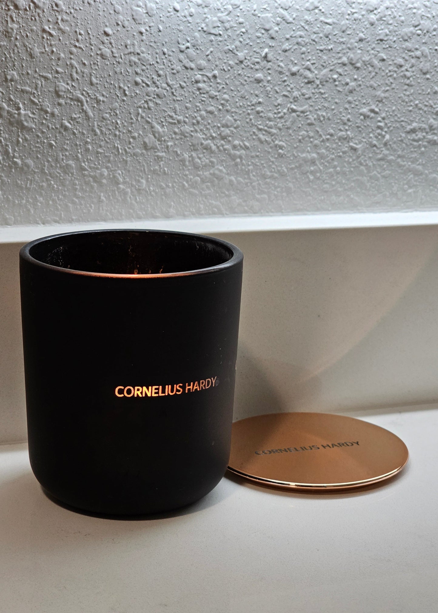 A black scented candle with an engraved jar, lit, showing the word 'CORNELIUS HARDY' glowing through the container, with a lid placed beside it on a white surface.