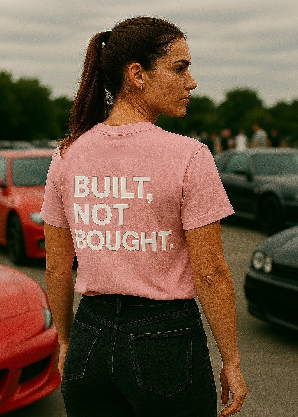 Woman wearing a pink t-shirt with 'BUILT, NOT BOUGHT.' text in an outdoor setting with cars.