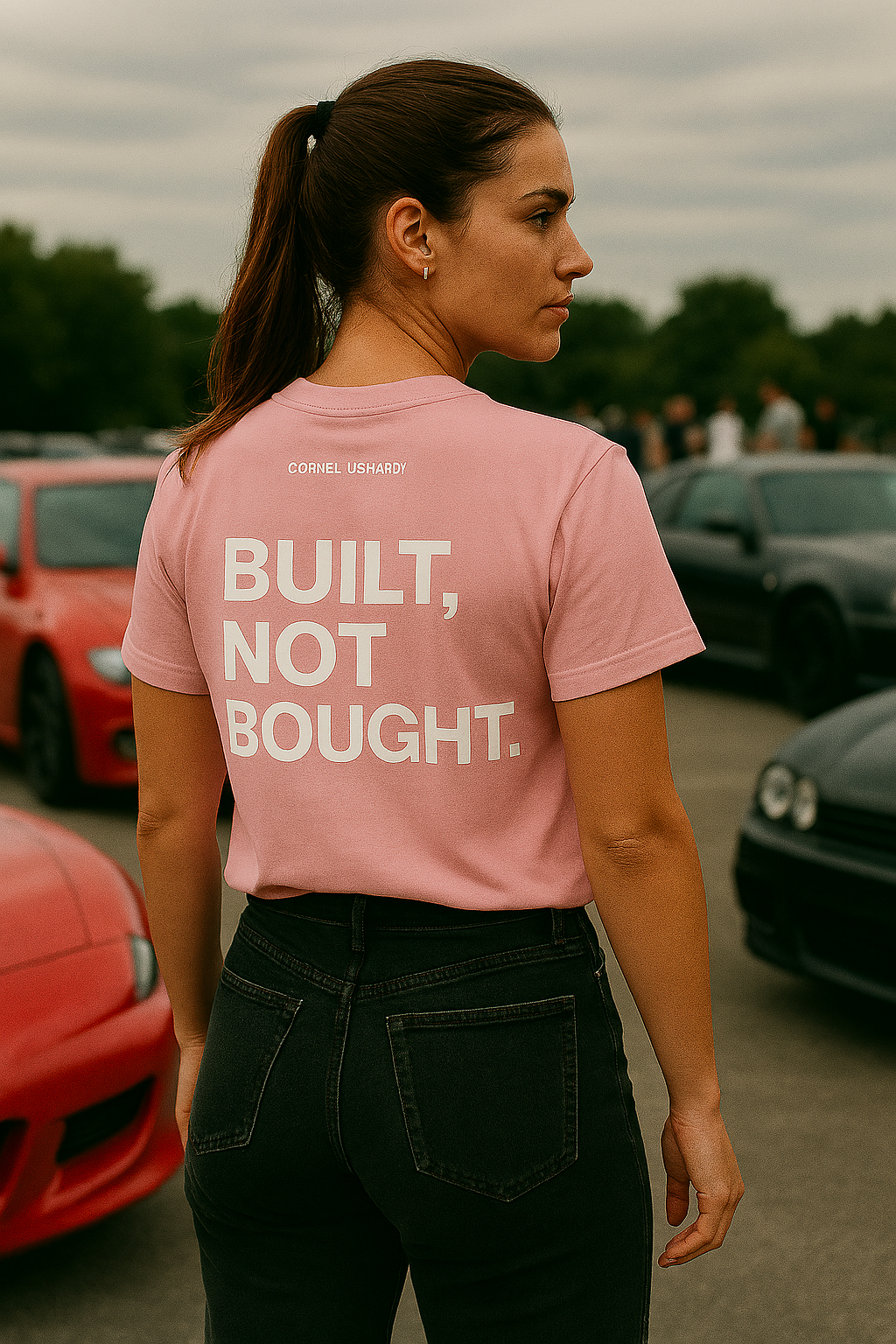 Woman wearing a pink t-shirt with 'BUILT, NOT BOUGHT.' text in an outdoor setting with cars.