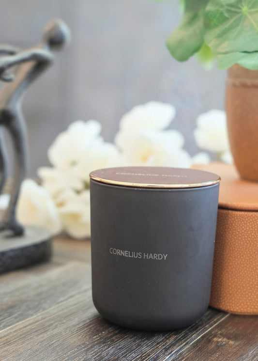 Black candle container with 'Cornelius Hardy' branding on a wooden surface with decorative items in the background.