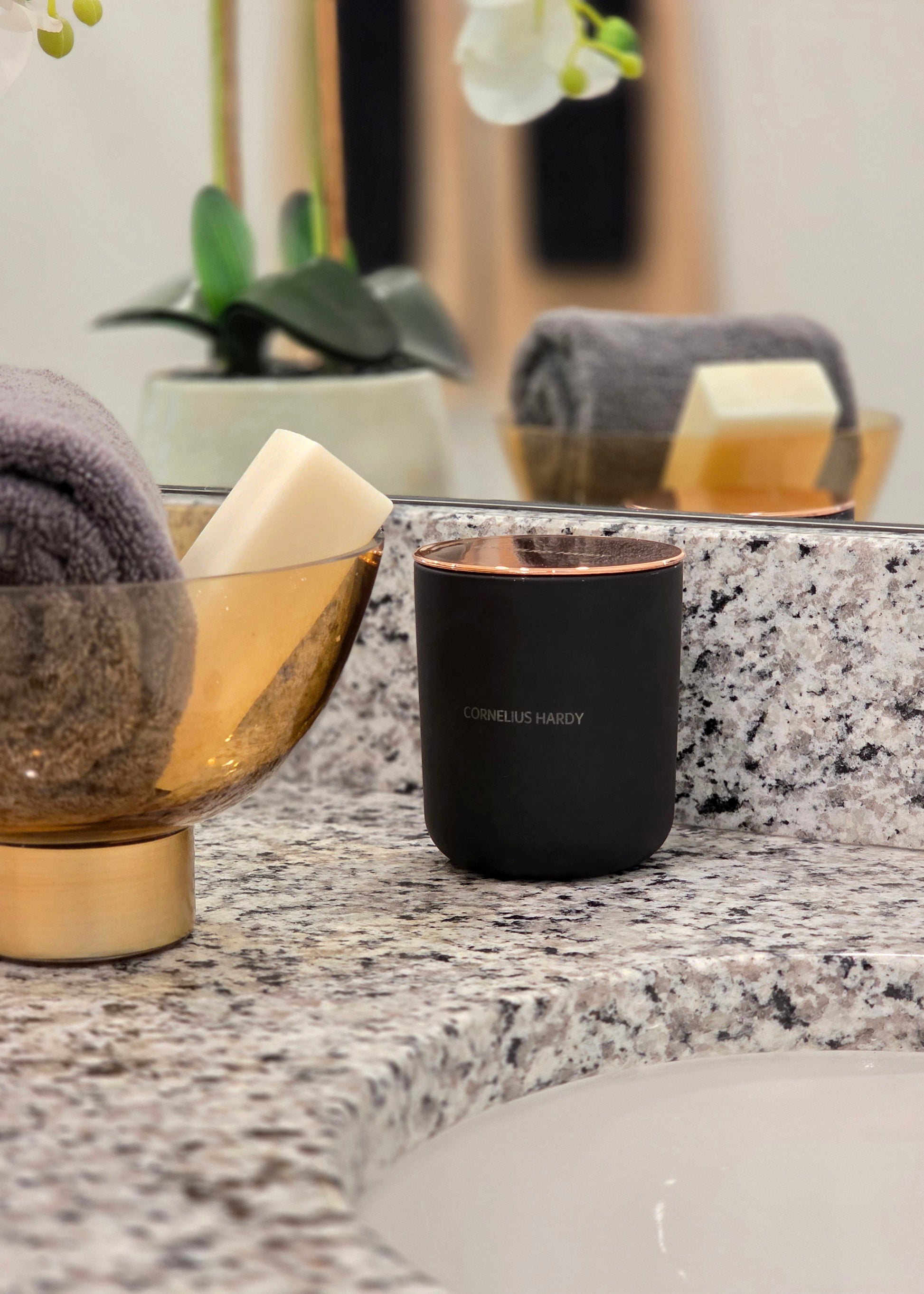 Black luxury candle on a bathroom counter with towels and soap in the background