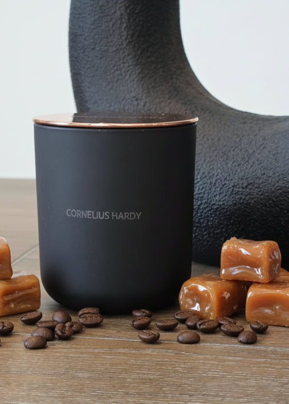 Black ceramic candle with 'Cornelius Hardy' branding on a wooden surface with a textured black object in the background.