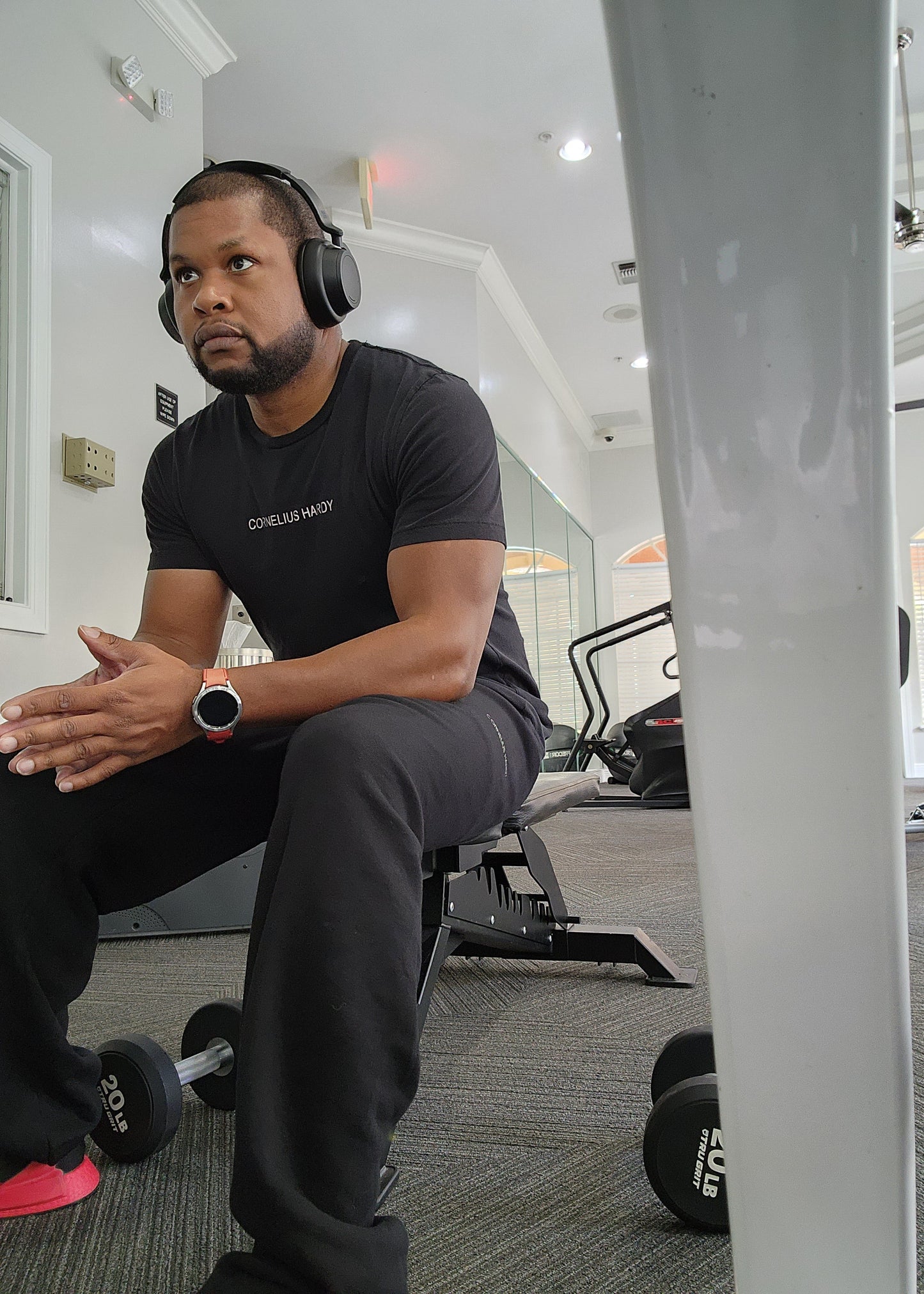 A man sitting in a gym wearing a black t-shirt with a logo on the chest, black pants, and headphones.