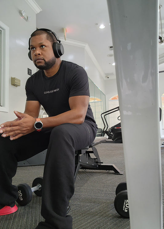 A man sitting in a gym wearing a black t-shirt with a logo on the chest, black pants, and headphones.
